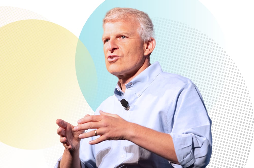 A middle-aged man with gray hair and glasses, wearing a light blue shirt, stands in front of a colorful abstract background.