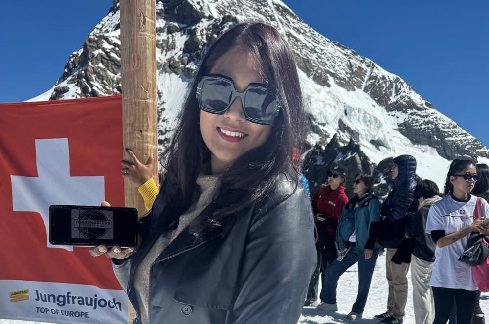 A smiling young woman holding a Swiss flag stands in front of a snowy mountain backdrop, surrounded by other people.