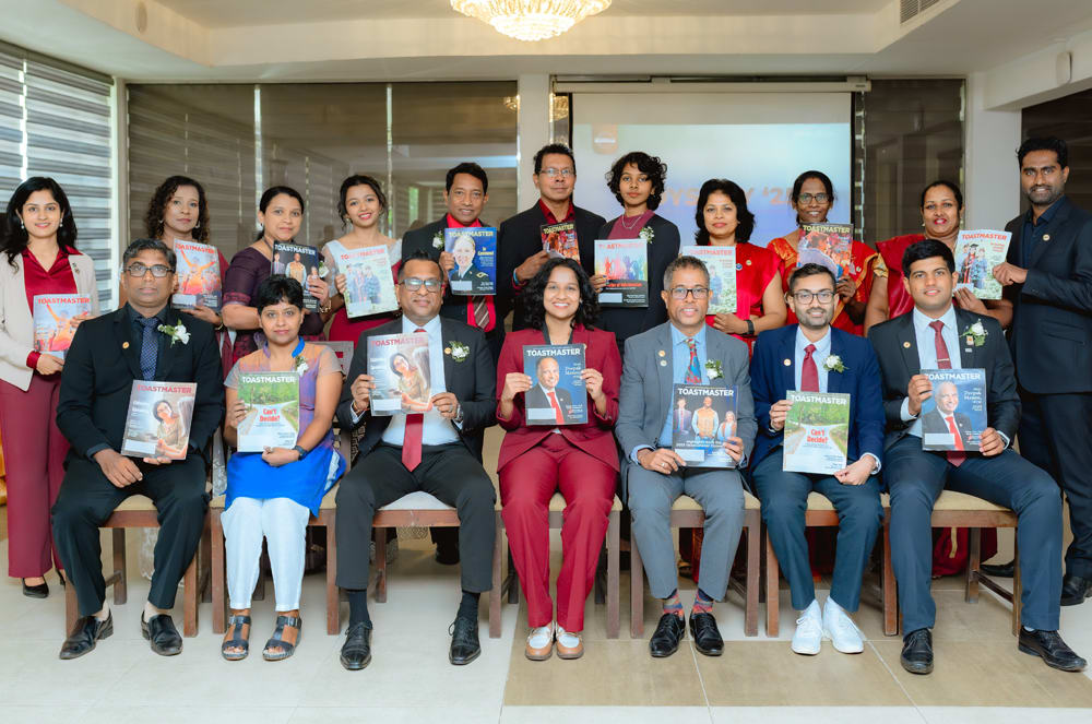 A group of people, likely students or graduates, are seated together holding certificates or diplomas, with a chandelier and other furnishings visible in the background.