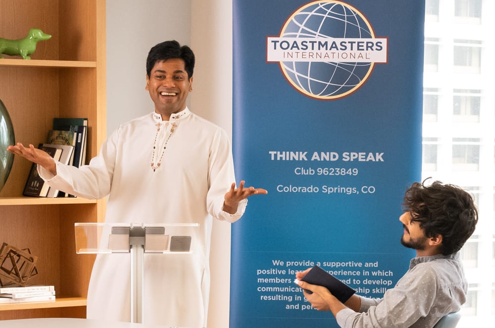 The image shows a man in a white shirt standing in front of a Toastmasters International banner, gesturing with his hands as he speaks to another person.