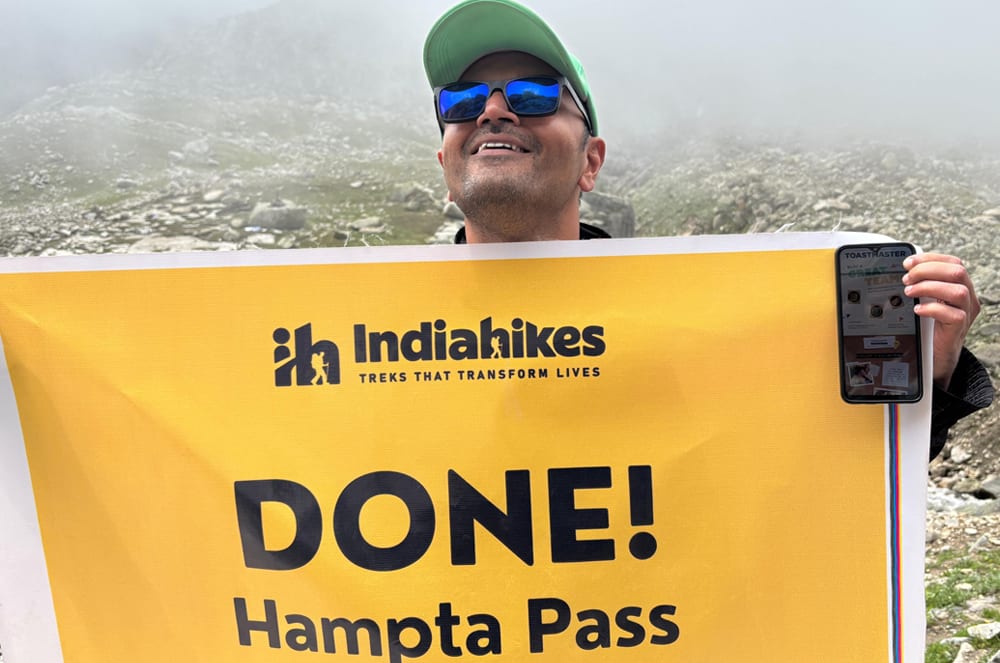 A person wearing sunglasses and a green hat holds a yellow sign that says "DONE! Hampta Pass" in front of a snowy mountain landscape.