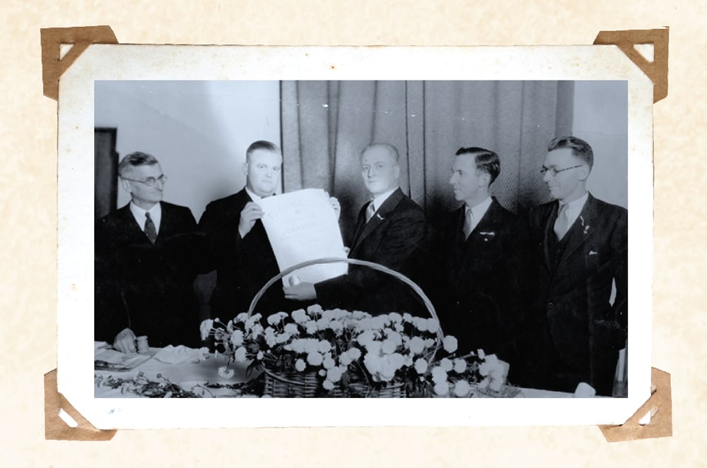 A group of men in suits standing around a table with a floral arrangement in the foreground.
