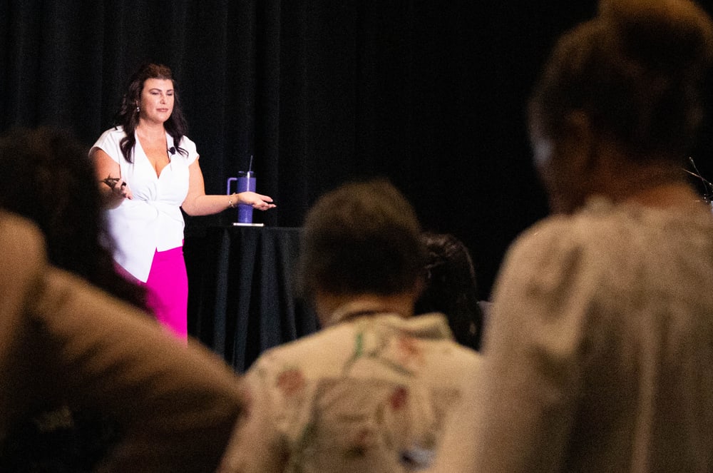 A woman in a white top and pink skirt is standing on a stage, addressing a group of people seated in the audience.