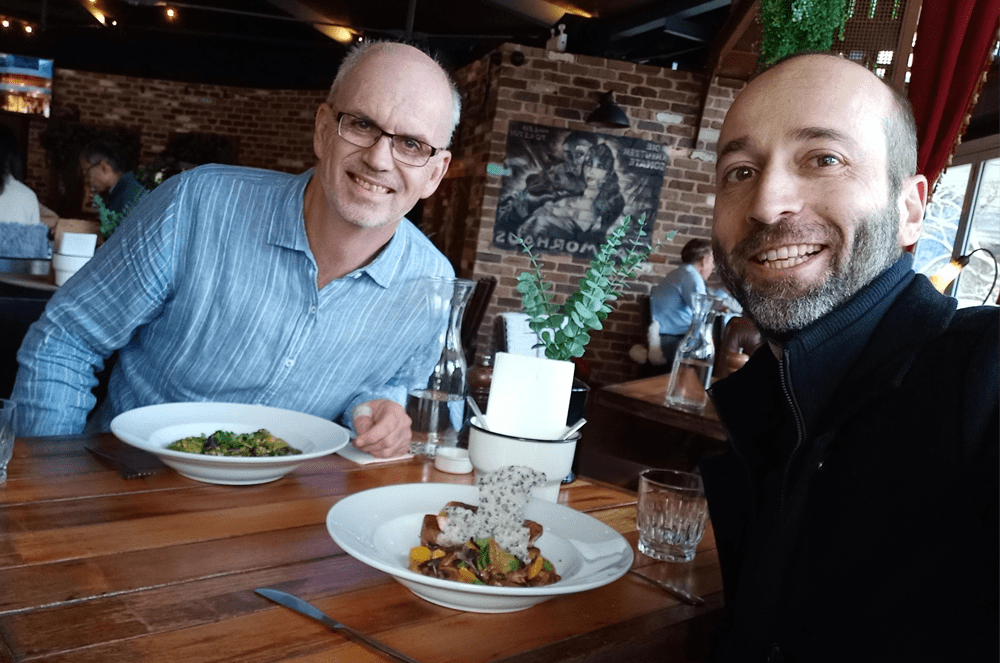 Two men sitting at a table in a restaurant, enjoying a meal and drinks. The background includes a brick wall with a framed artwork and other decor elements.