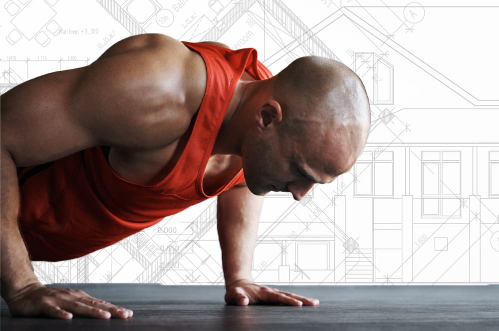 A muscular man in a red tank top is performing a push-up on a wooden floor, with architectural drawings visible in the background.
