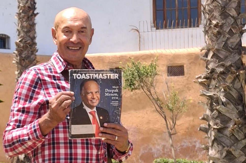 A smiling older man in a plaid shirt holding a Toastmaster magazine in front of a building with palm trees in the background.