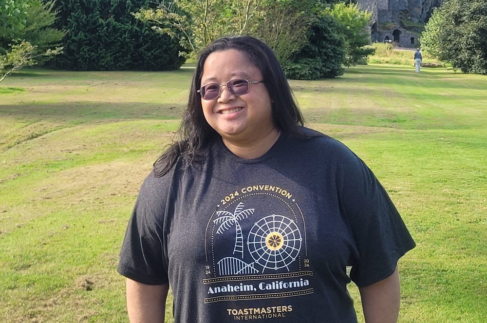 A smiling person wearing a black t-shirt with a graphic design stands in a grassy outdoor setting with trees and buildings visible in the background.