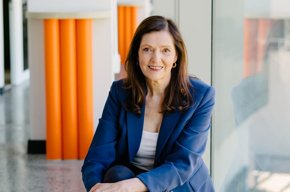 A smiling woman in a blue blazer stands in front of a bright orange background.