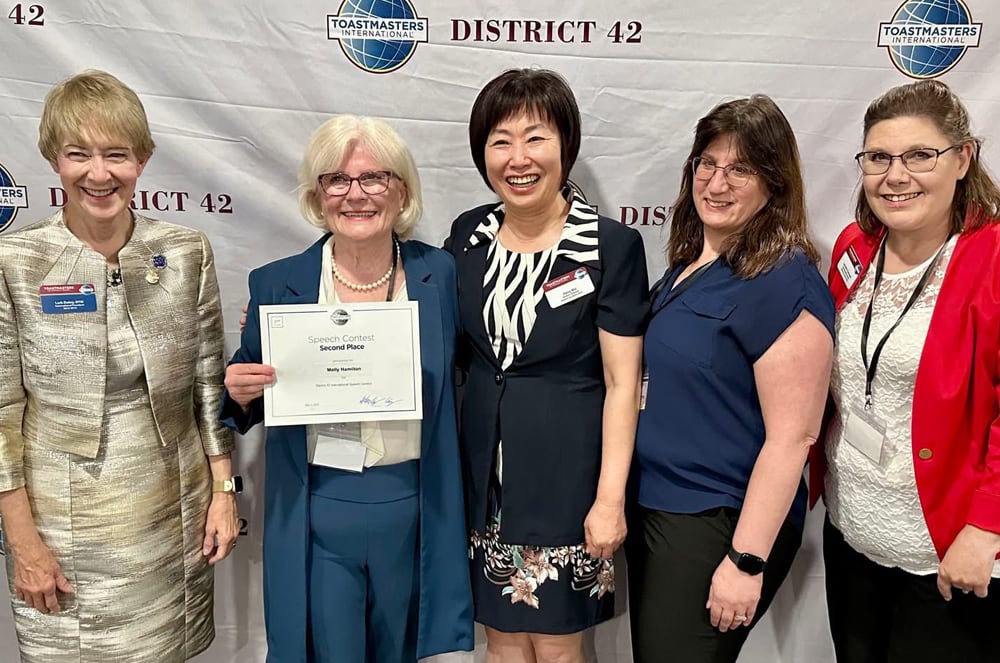 The image shows a group of five women standing together in front of a banner for "District 42", with each woman holding a certificate or award.