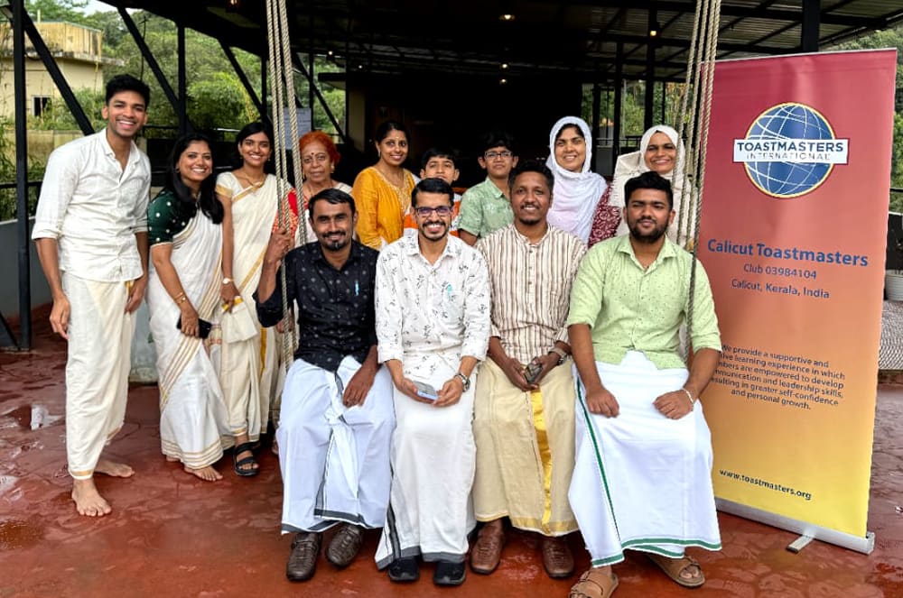 A group of people, both men and women, are standing together in front of a Toastmasters banner, dressed in a variety of casual and formal attire, suggesting a community or organizational event.