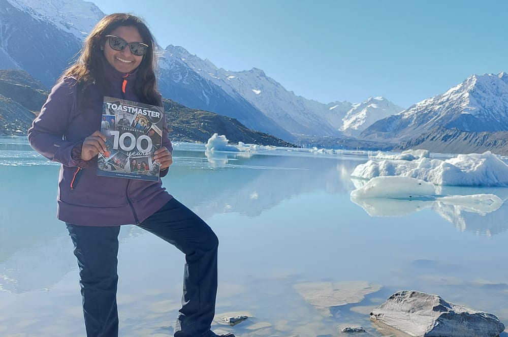 A smiling woman holding the Toastmaster magazine near a glacier
