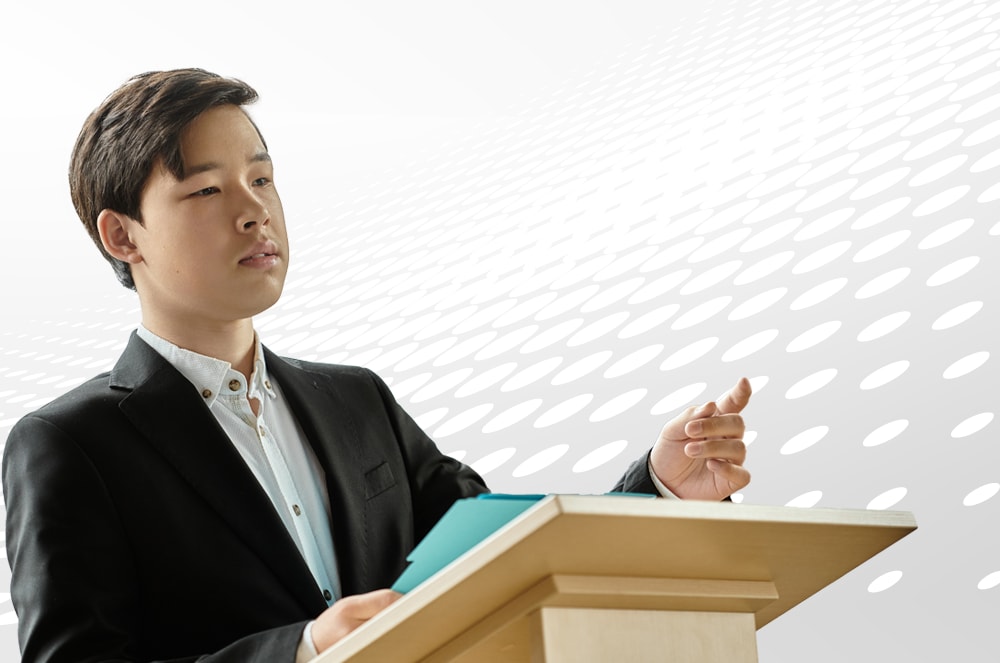 A teenager in a black suit jacket stands at a lectern