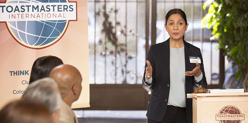 The image shows a woman in a black blazer standing at a podium with the Toastmasters International logo, while an audience is visible in the background.