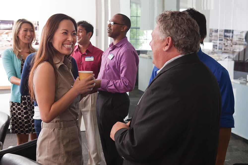 A group of people, some in casual attire and others in professional business wear, are engaged in conversation and interaction in what appears to be a professional or office setting.