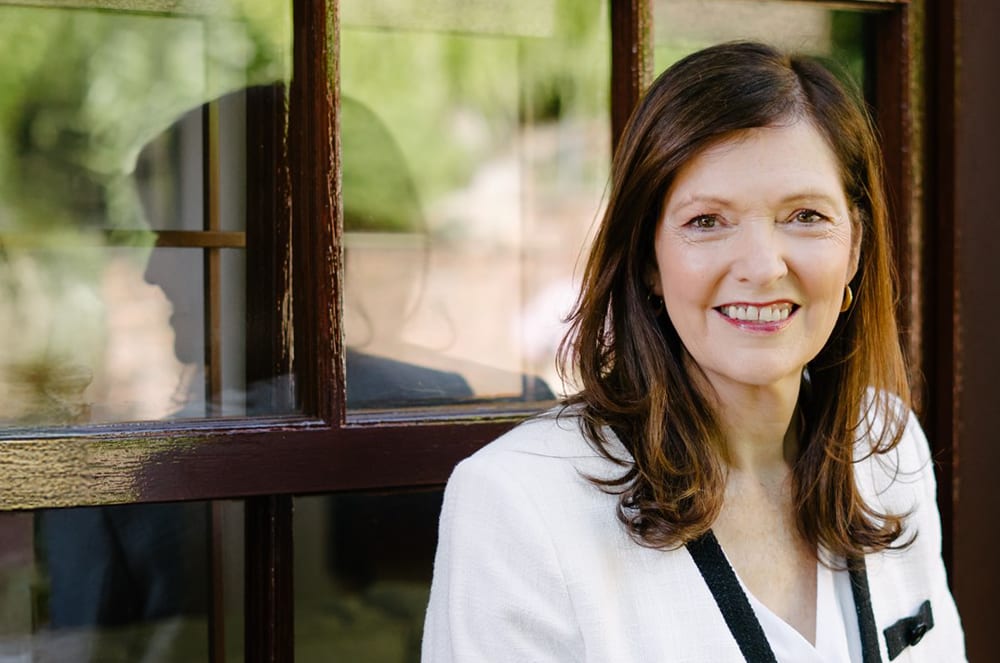 A smiling woman with long brown hair stands in front of a wooden window frame, with a blurred natural background visible behind her.