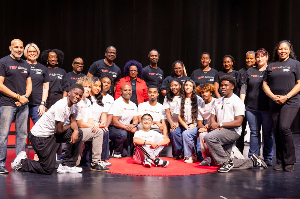 A group of people, both adults and children, are gathered on a stage, wearing various t-shirts and posing for a photograph.