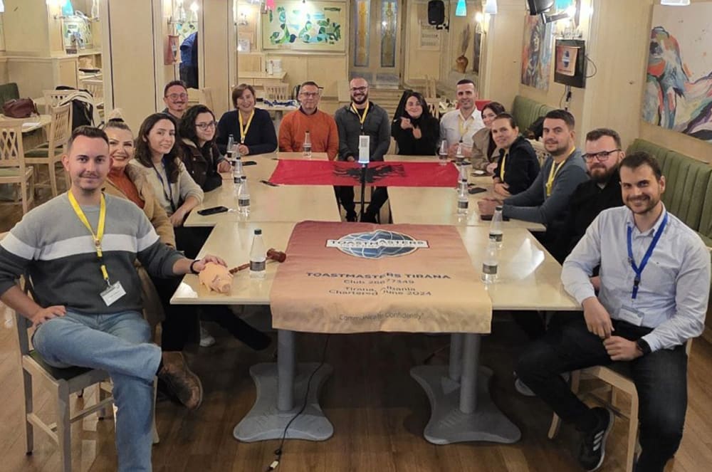 A group of people gathered around a table, some sitting and some standing, in what appears to be a casual meeting or gathering in a cozy, decorated indoor space.