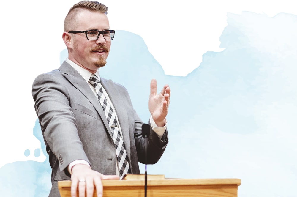 A man in a suit and glasses stands behind a podium, gesturing with his hands as he speaks.