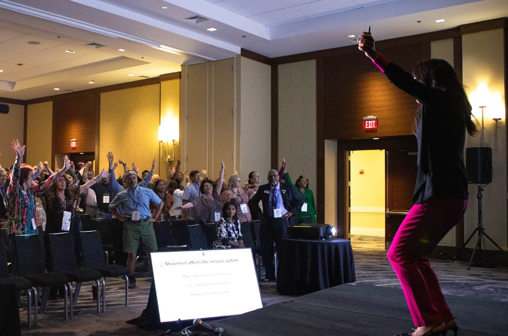 A large group of people gathered in a conference room, with a person standing on a stage gesturing to the audience.