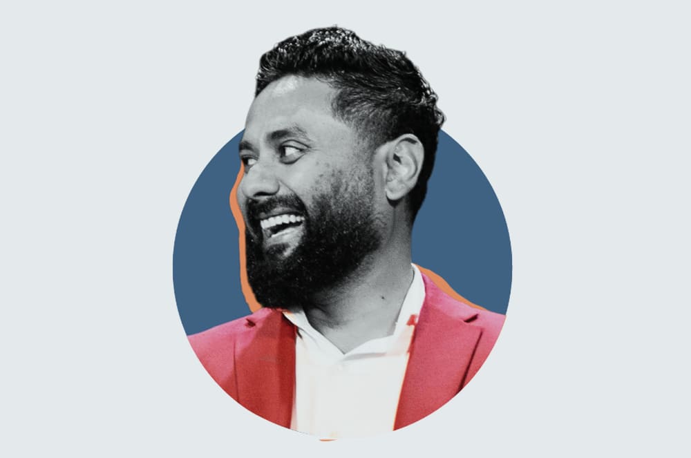 A smiling man with a beard wearing a pink jacket against a blue background.