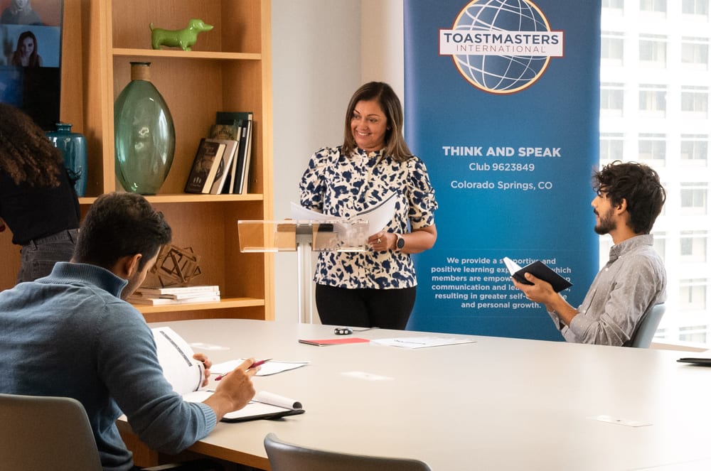 A woman in a patterned blouse is standing in front of a Toastmasters banner, addressing a group of people seated around a table.
