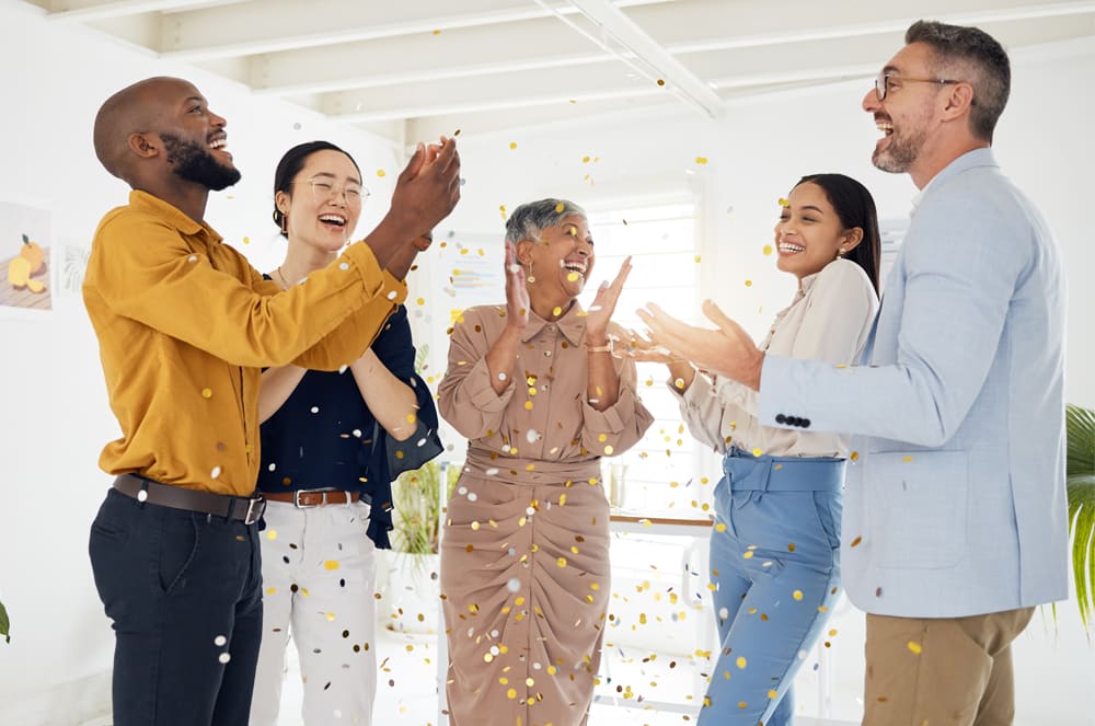 A group of five people, three women and two men, are standing together and celebrating with confetti in a bright, open room.