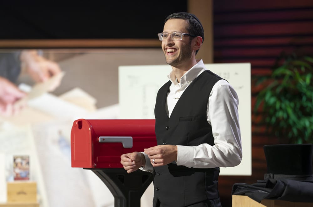 A man in a suit and glasses stands in front of a red mailbox, with a backdrop of a room with plants and other office-like furnishings.