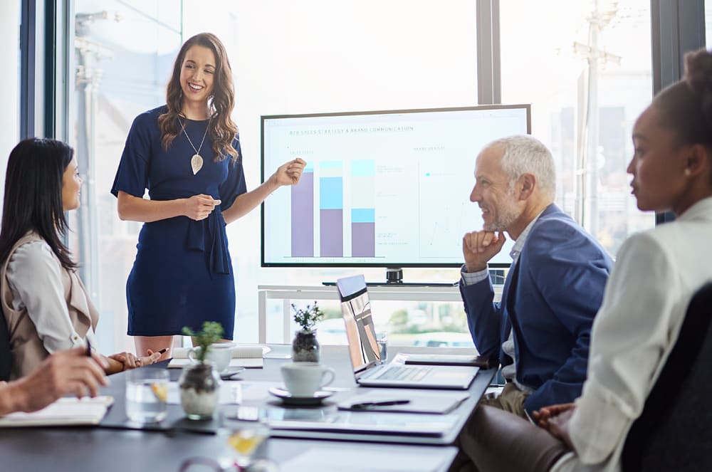 A smiling man in a light blue shirt stands in front of a whiteboard displaying various charts and graphs.