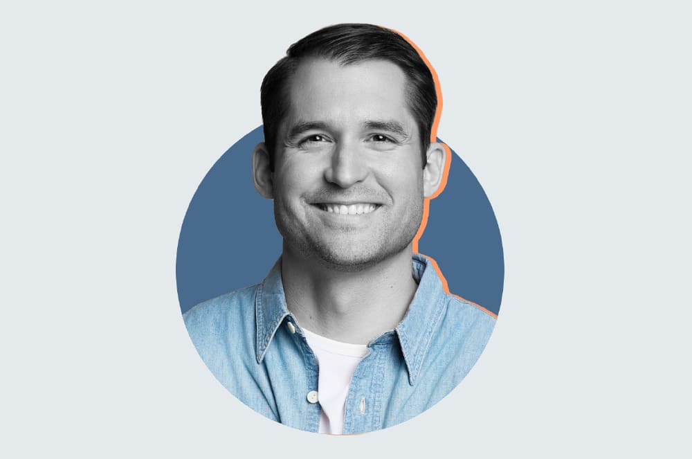 A smiling man with a friendly expression, wearing a light blue denim shirt, set against a plain blue background.