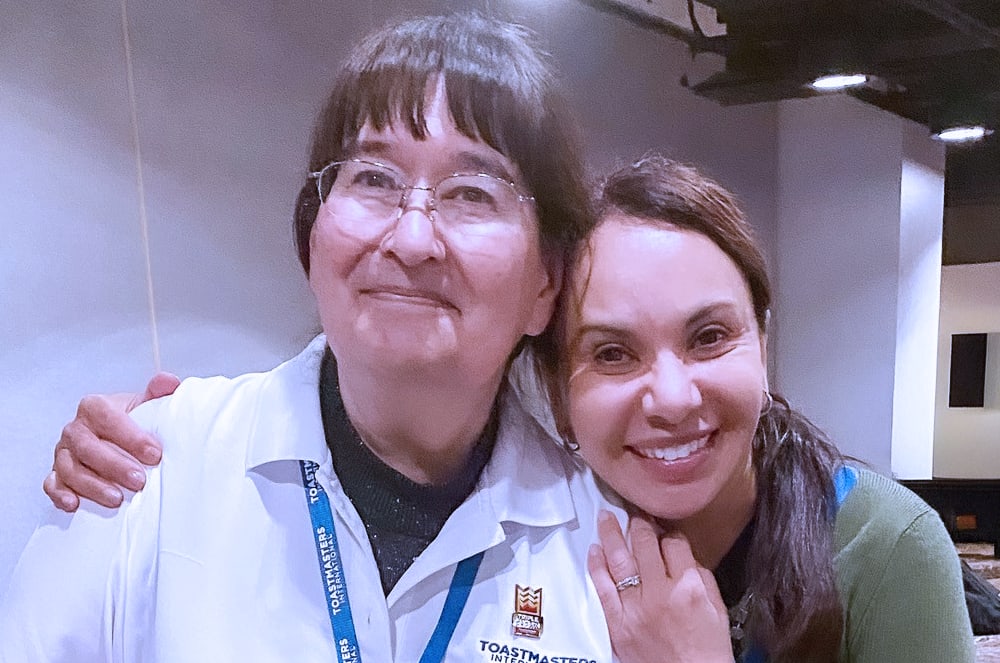 The image shows two women, an older woman with glasses and a younger woman, embracing and smiling warmly in what appears to be a professional or work setting.