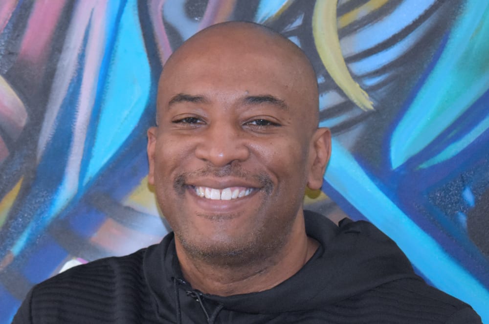 A smiling man with a bald head stands in front of a vibrant, colorful mural backdrop.