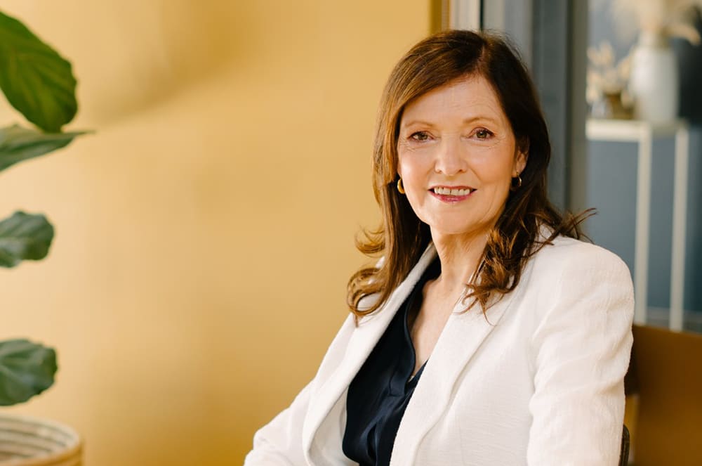 A smiling woman with curly brown hair wearing a white blazer stands in front of a light-colored wall with a potted plant visible in the background.