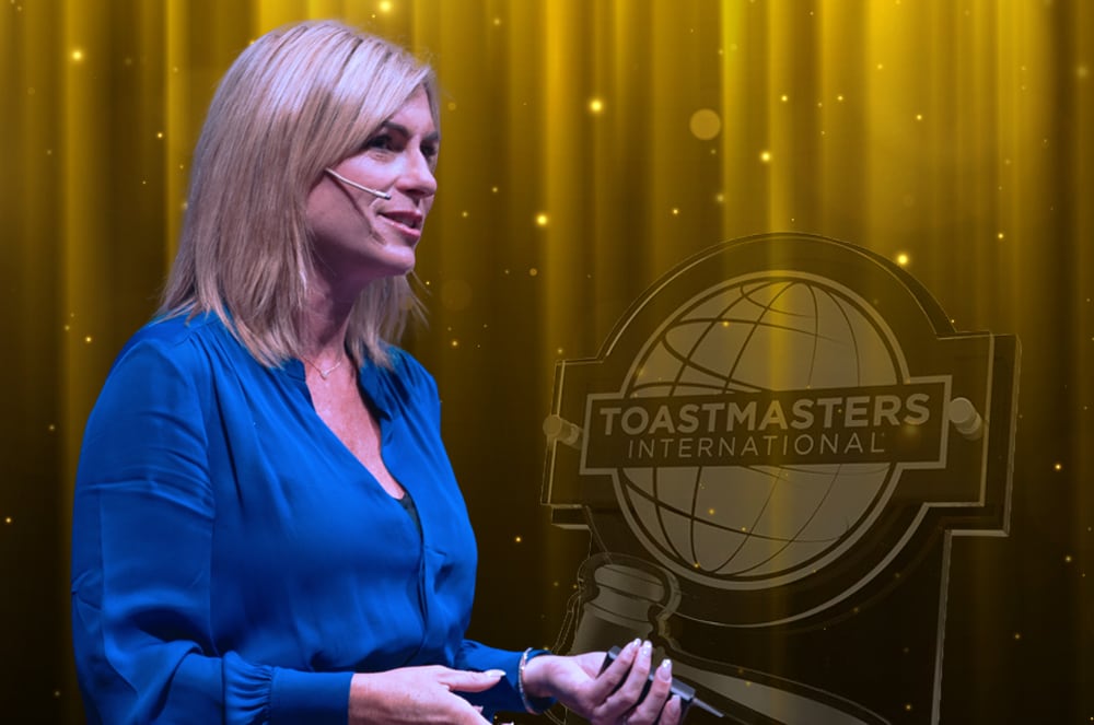 A woman with blonde hair stands in front of a Toastmasters International backdrop, wearing a blue blouse and smiling.