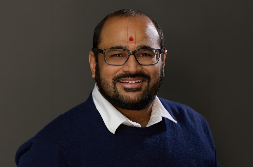 A smiling man with glasses and a bindi on his forehead, wearing a navy blue sweater, stands against a dark background.