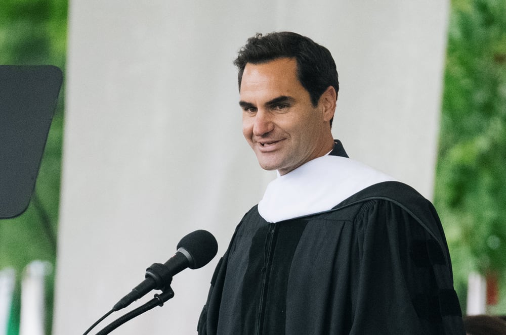 The image shows a man in a black academic gown standing at a podium and speaking into a microphone, with a green background behind him.