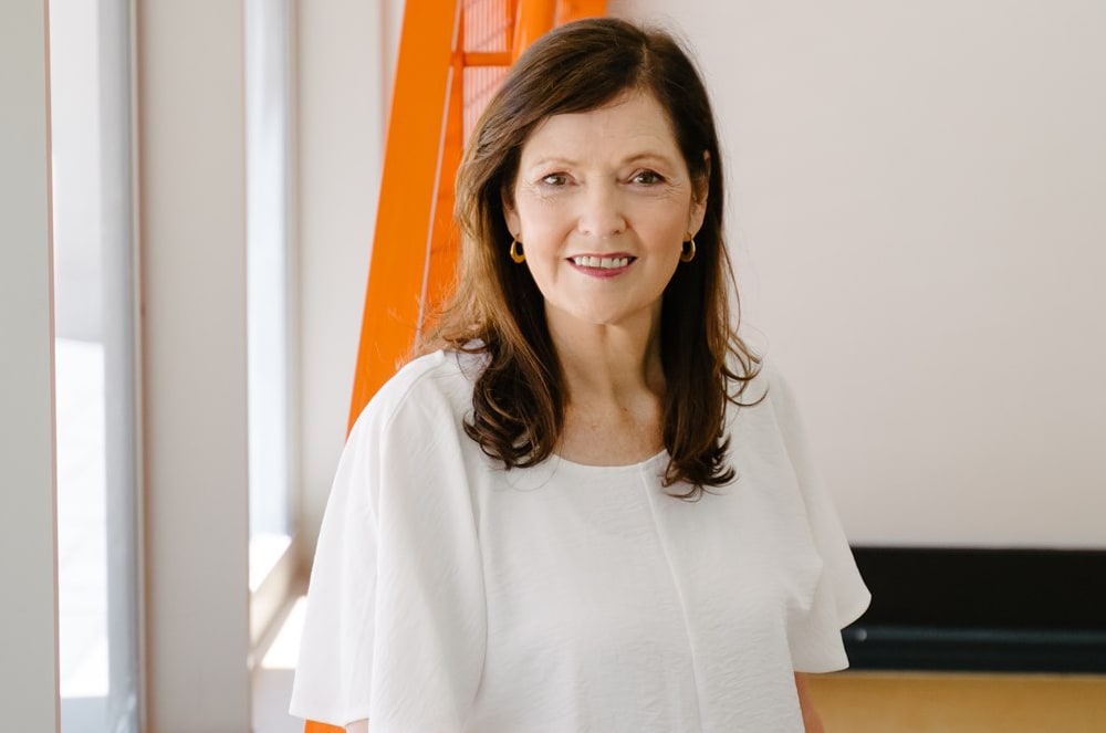 A smiling woman with long brown hair stands in front of an orange window frame, creating a warm and inviting atmosphere.