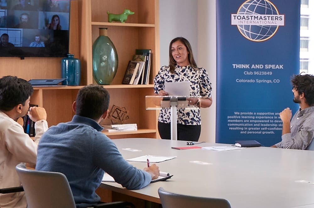 A woman in a patterned blouse is standing at a podium in front of a Toastmasters banner, addressing a group of people seated around a table.