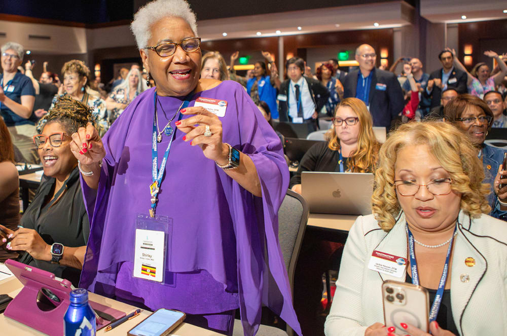 A smiling elderly woman in a purple robe stands in a crowded room filled with people, some of whom are also wearing conference badges.