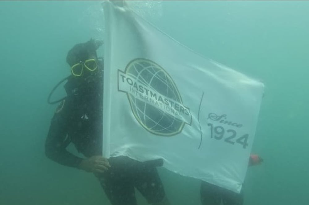 A diver holding a large white flag with the Toastmasters International logo underwater in a green-tinted body of water.