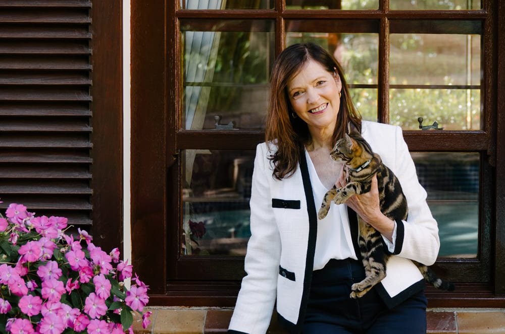 A woman in a white blazer smiles while holding a tabby cat in front of a window with pink flowers to the left.