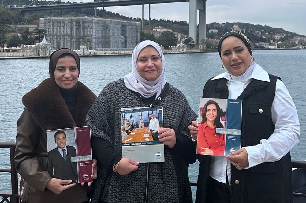 Three women stand on a deck holding brochures, with a large bridge and buildings visible across a body of water in the background.