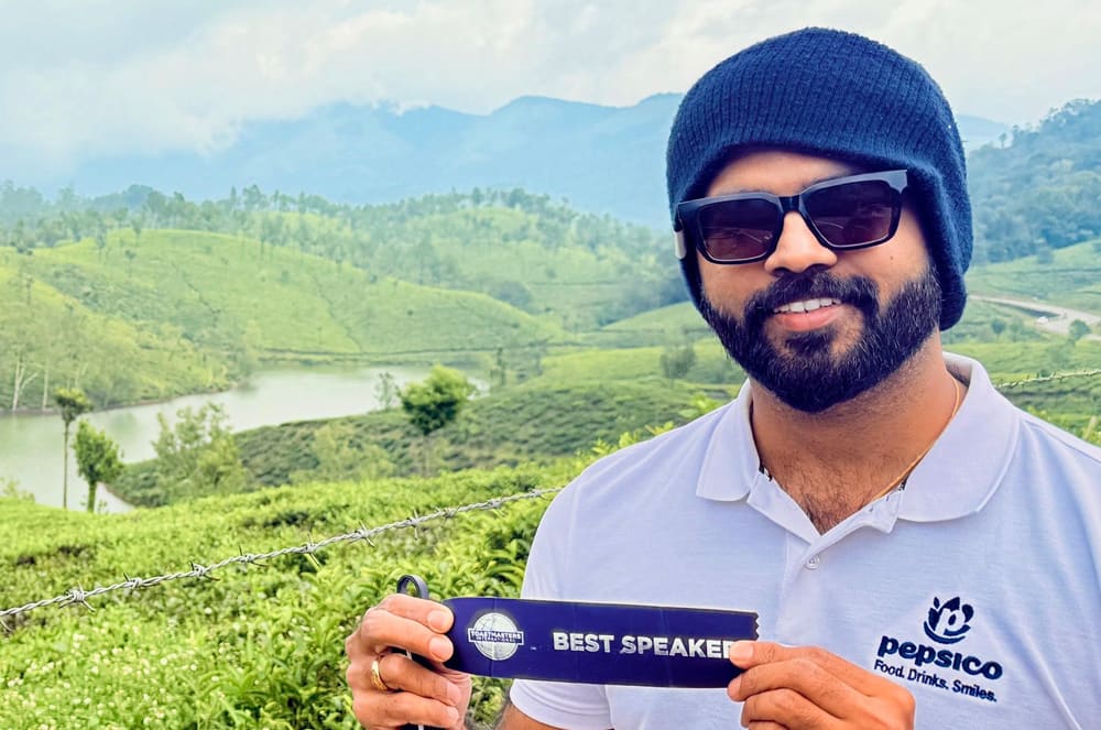 A man in a beanie and sunglasses smiles at the camera, holding a "Best Speaker" ribbon in front of a lush green landscape with rolling hills and a body of water.