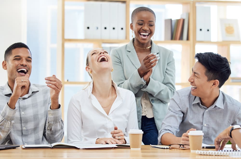 Four colleagues laugh heartily around a table in the foreground, with shelves of books and office supplies visible in the blurred background.