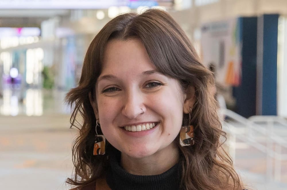 A young woman with brown hair and a nose ring smiles at the camera in the foreground, with a blurred, brightly lit indoor space in the background.