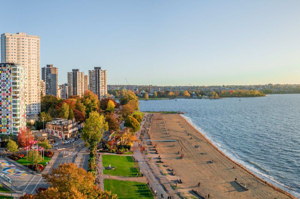 A sandy beach with scattered people stretches along a waterfront park with colorful autumn trees, backed by a city skyline of tall buildings and a calm blue bay under a clear sky.
