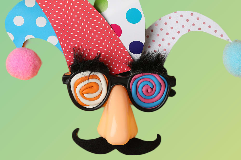 A jester's hat and silly glasses with a clown nose and mustache are in the foreground against a soft green background.
