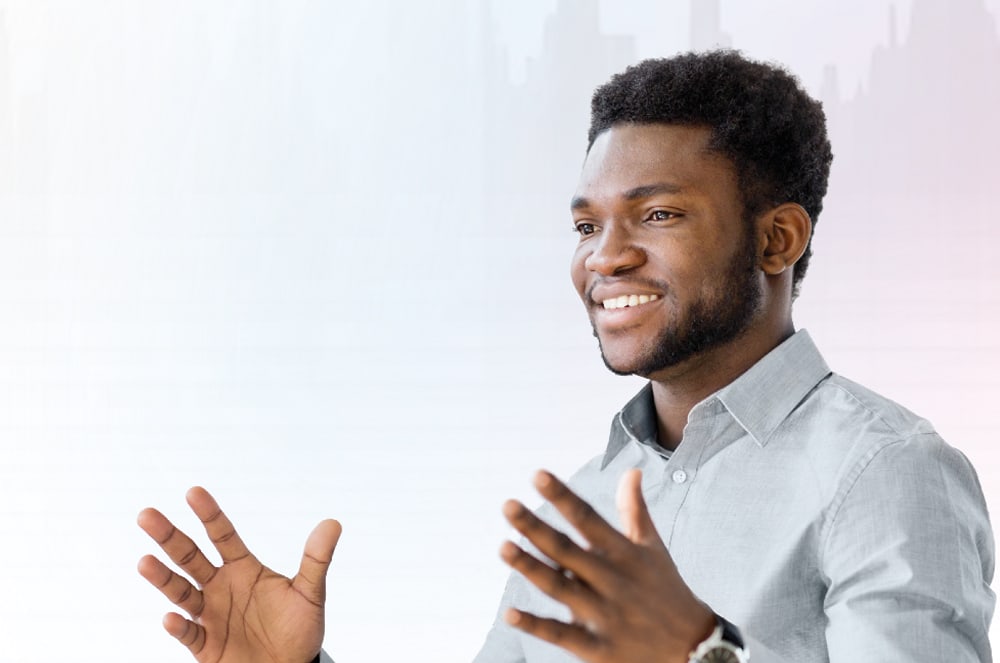 A smiling Black man in a light blue collared shirt gestures with his hands in the foreground, while a blurred, light-colored background with faint vertical lines suggests a professional setting.