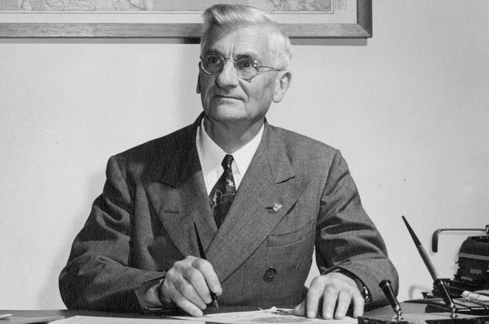 An elderly man in a suit sits at a desk with papers and a typewriter, looking off to the side with a framed map on the wall behind him.