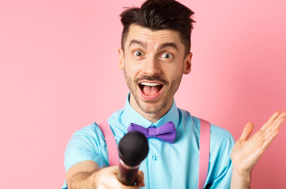 A man with a purple bowtie and pink suspenders holds a microphone forward against a pink background.