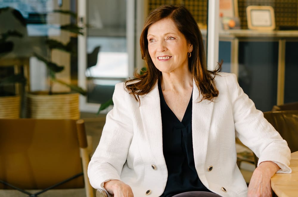 A woman in a white blazer smiles while looking off to the side, with blurred office plants and furniture in the background.